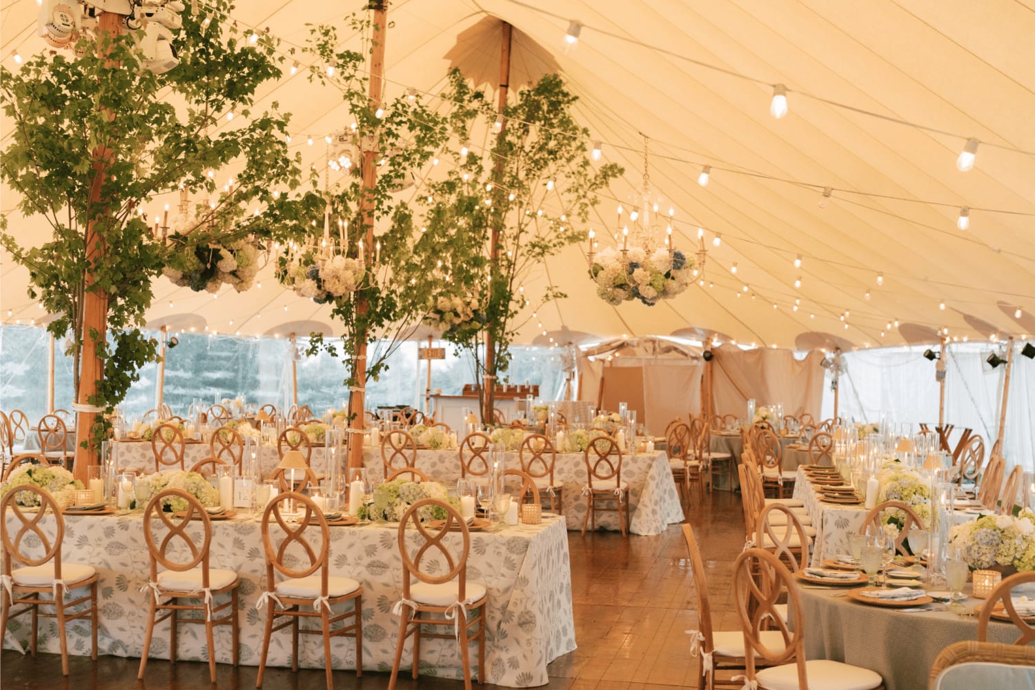 Photo of a wedding reception space, in a tent with floral chandelier installations, with floral centerpieces and candles on a mixture of round and farm tables.