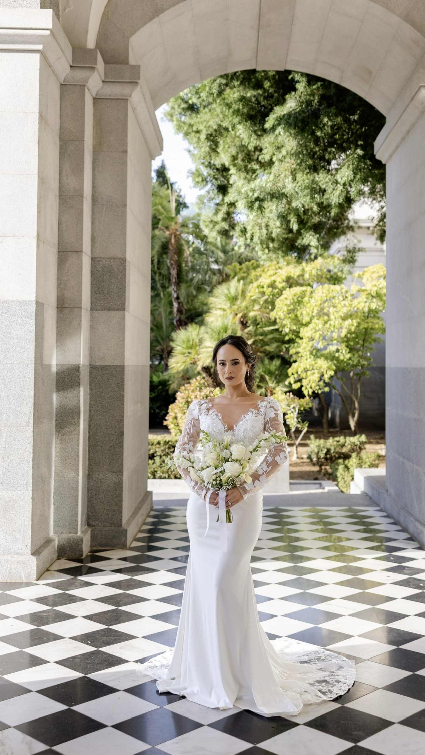 Bride in a white wedding gown holding an all-white floral bouquet beneath a stone archway.