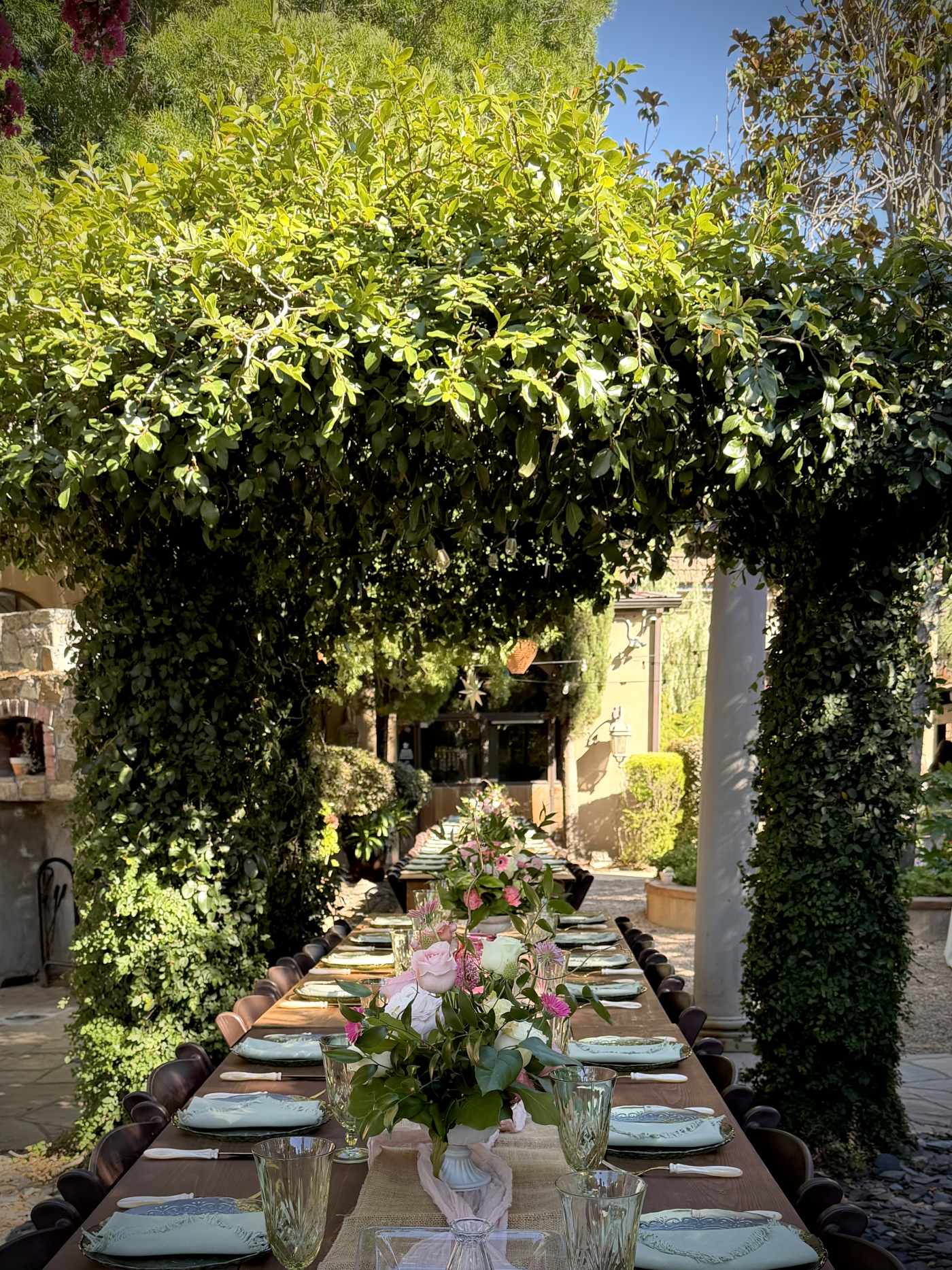 Long outdoor wedding table beneath a greenery-covered arch with pink and ivory floral centerpieces.