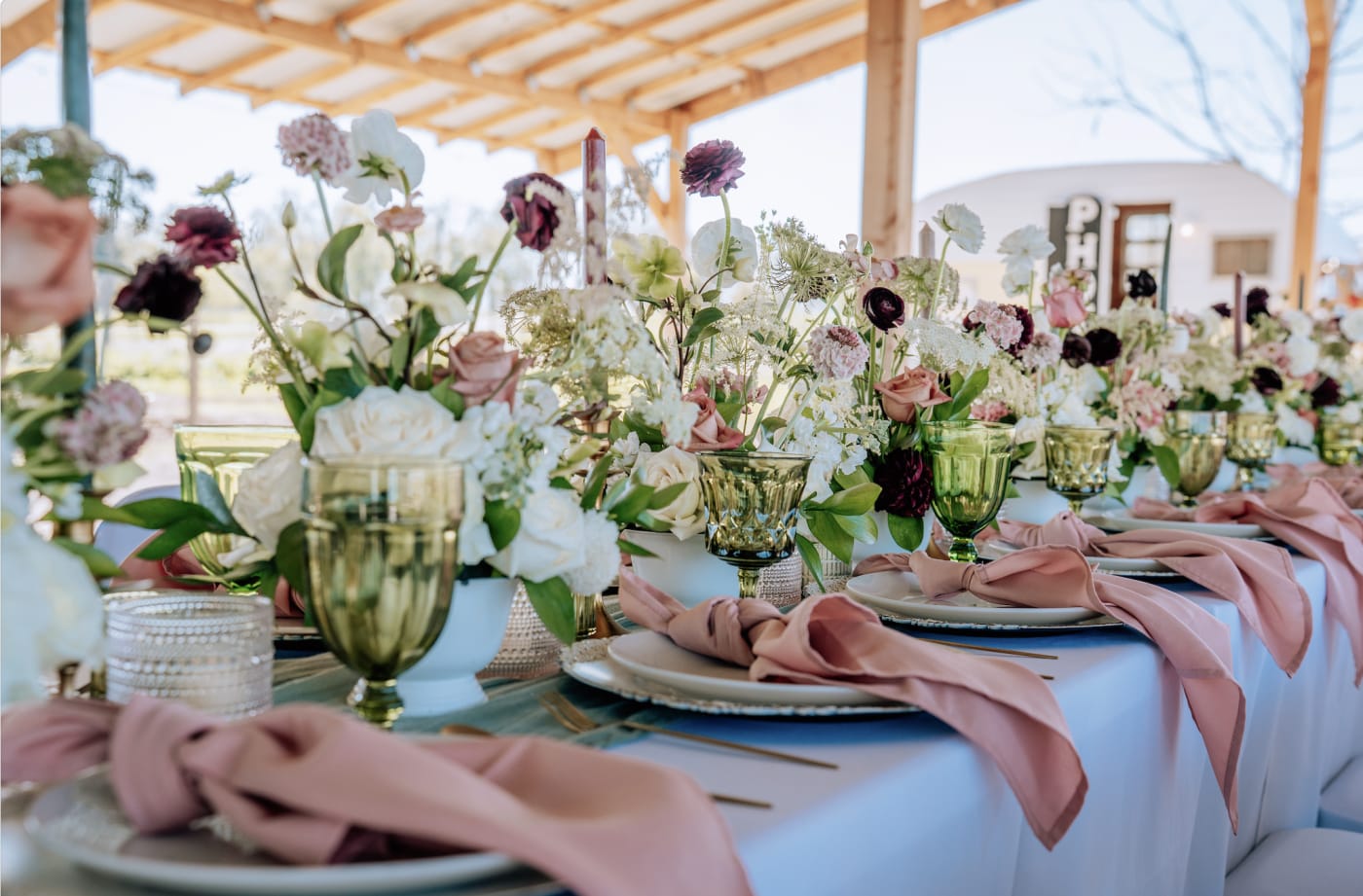 Elegant wedding reception table with airy pink, ivory and burgundy floral centerpieces, blush napkins and green glassware.