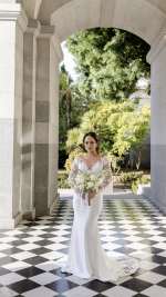 Bride in a white wedding gown holding an all-white floral bouquet beneath a stone archway.