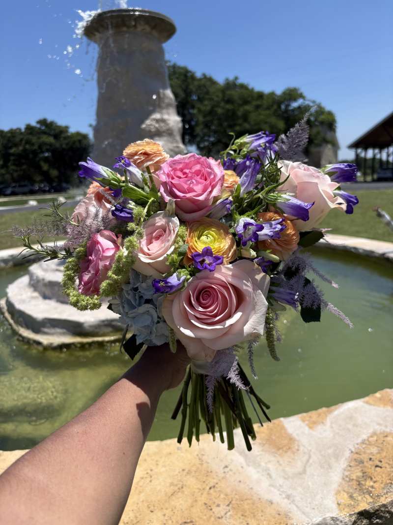 Featuring bubblegum pink roses, lavender campanula, peach ranunculus, soft blue hydrangea, and light pink tinted plumosa, the bouquet creates a fresh, garden-inspired look bursting with personality.