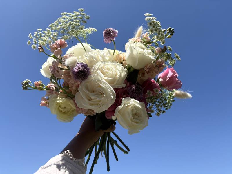 Featuring vibrant hot pink hydrangea, peach stock, lavender scabiosa, white roses, country blue roses, and delicate Queen Anne's lace, the arrangement feels both whimsical and refined.