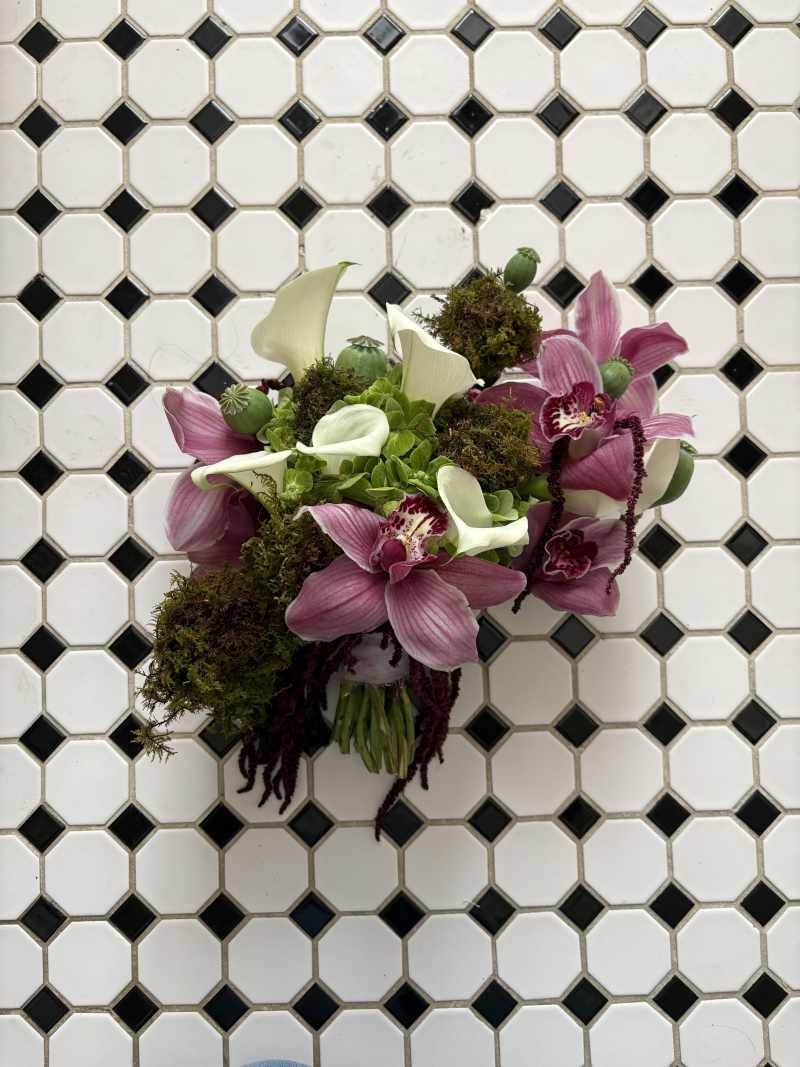 Featuring green hydrangeas, elegant white calla lilies, rich burgundy cymbidium orchids, cascading burgundy amaranthus, textured green poppy pods, and a dramatic moss sphere that flows into the bouquet's design.