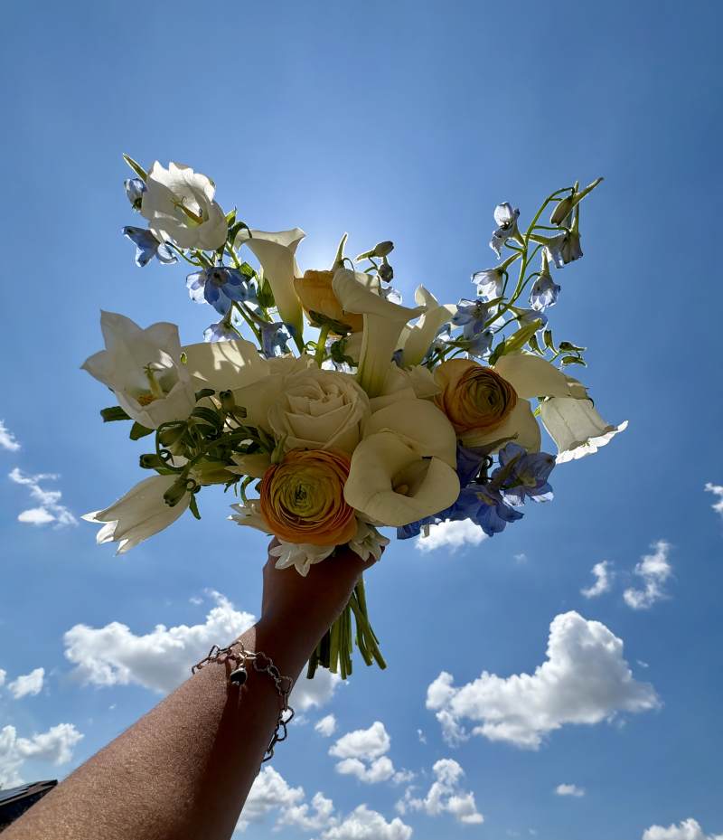 Featuring sculptural white calla lilies, delicate white campanula, light blue delphinium, classic white roses, and peach ranunculus, the bouquet creates a graceful and airy design full of movement and charm.
