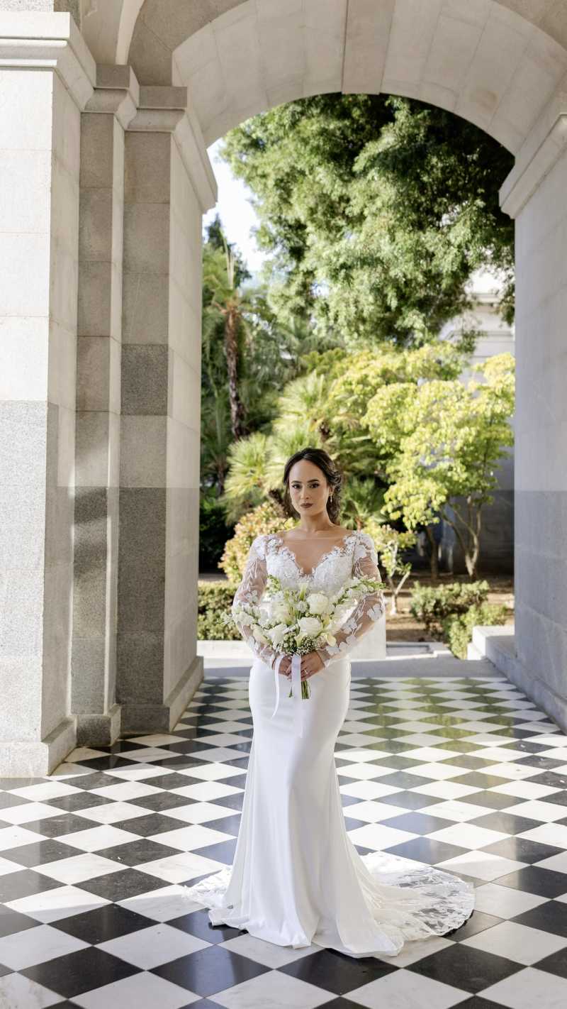Bride in a white wedding gown holding an all-white floral bouquet beneath a stone archway.