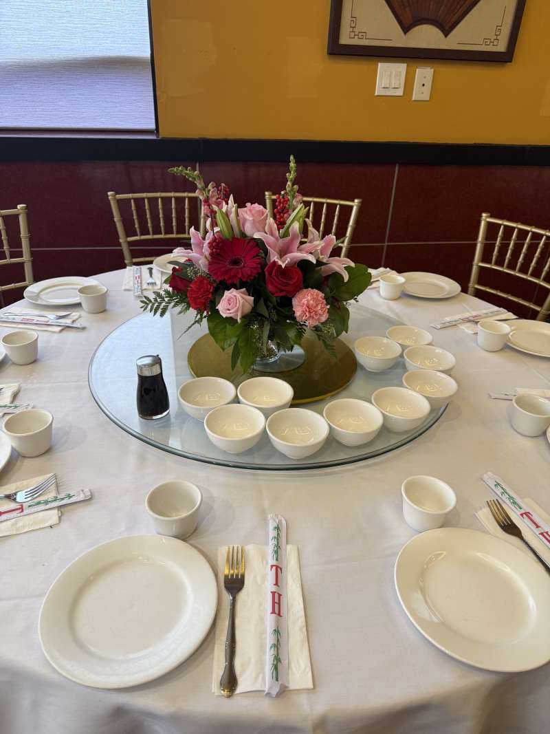 Reception table set with white linens, gold chairs, and a red and pink floral centerpiece featuring roses, lilies, and greenery on a glass lazy Susan.
