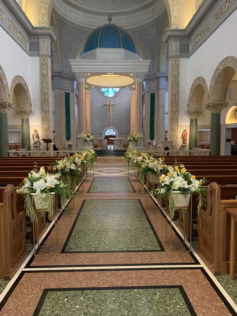 Wedding ceremony at The Immaculata Parish on the University of San Diego campus featuring wooden pews, elevated aisle floral arrangements in white and yellow, and elegant church architecture.