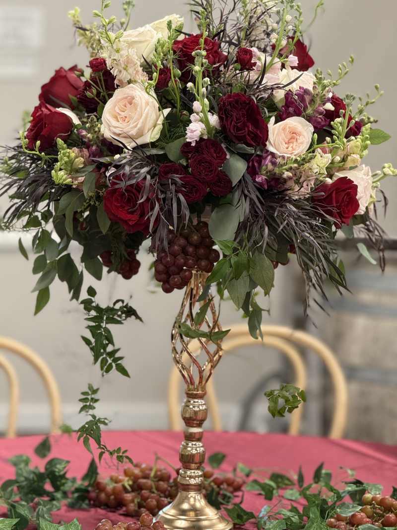 Tall wedding centerpiece with burgundy and blush flowers, greenery, and grapes on a gold stand.