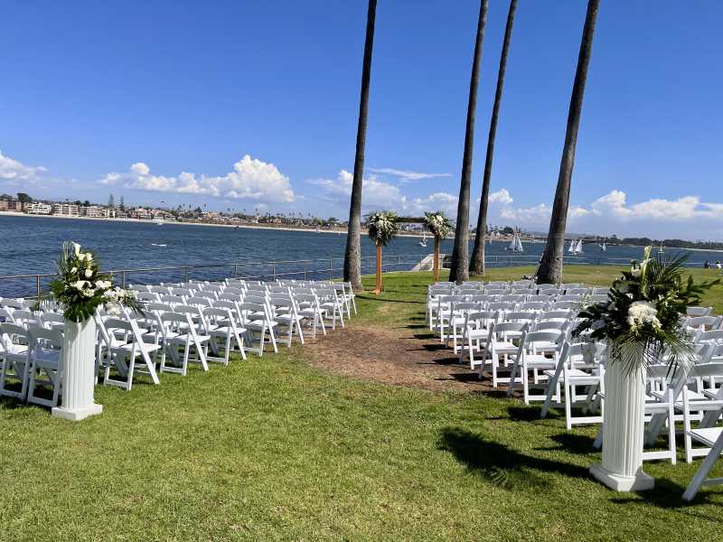 Outdoor waterfront wedding ceremony at Tower Beach Club featuring rows of white chairs, tropical white and green floral arrangements, palm trees, and a wooden arch overlooking the water in San Diego.