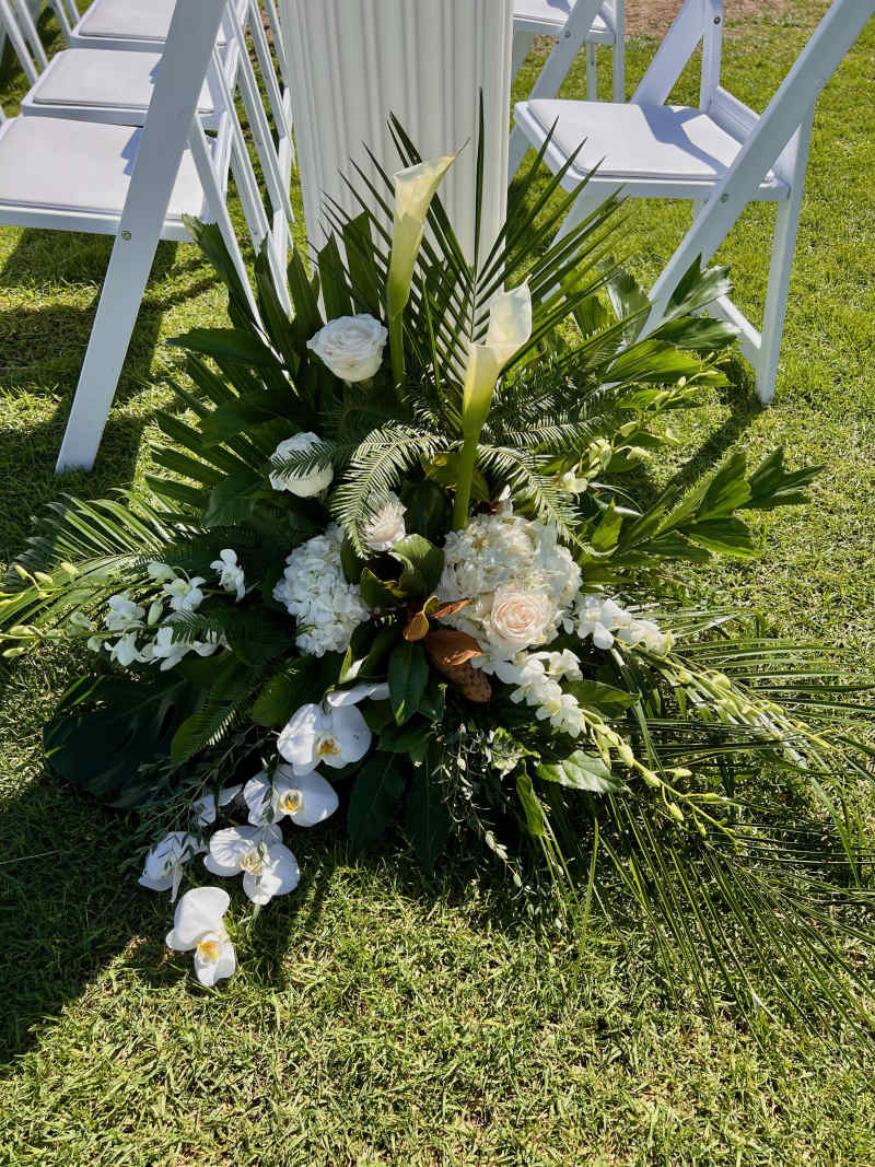 Lush grounded floral arrangement with white calla lilies, orchids, roses, hydrangeas, and tropical greenery styled beside ceremony chairs for an aisle marker or entryway design.