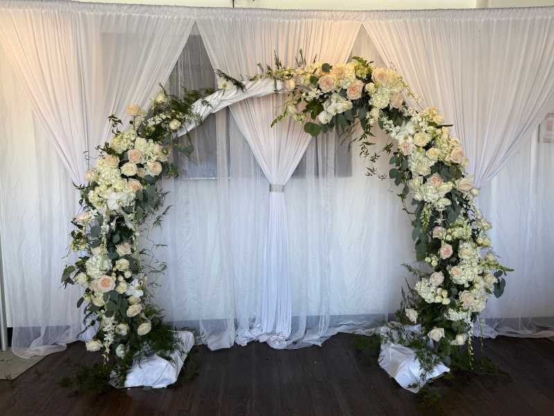 Romantic circular wedding arch covered in white roses, hydrangeas, orchids, and greenery, styled with white draping and a soft fabric backdrop.
