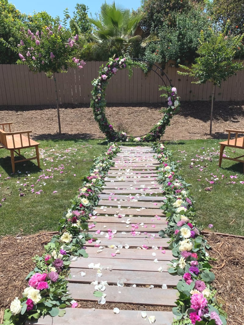 Outdoor backyard wedding ceremony at a private residence featuring a wooden aisle lined with pink, purple, and white flowers, scattered rose petals, and a circular floral arch.