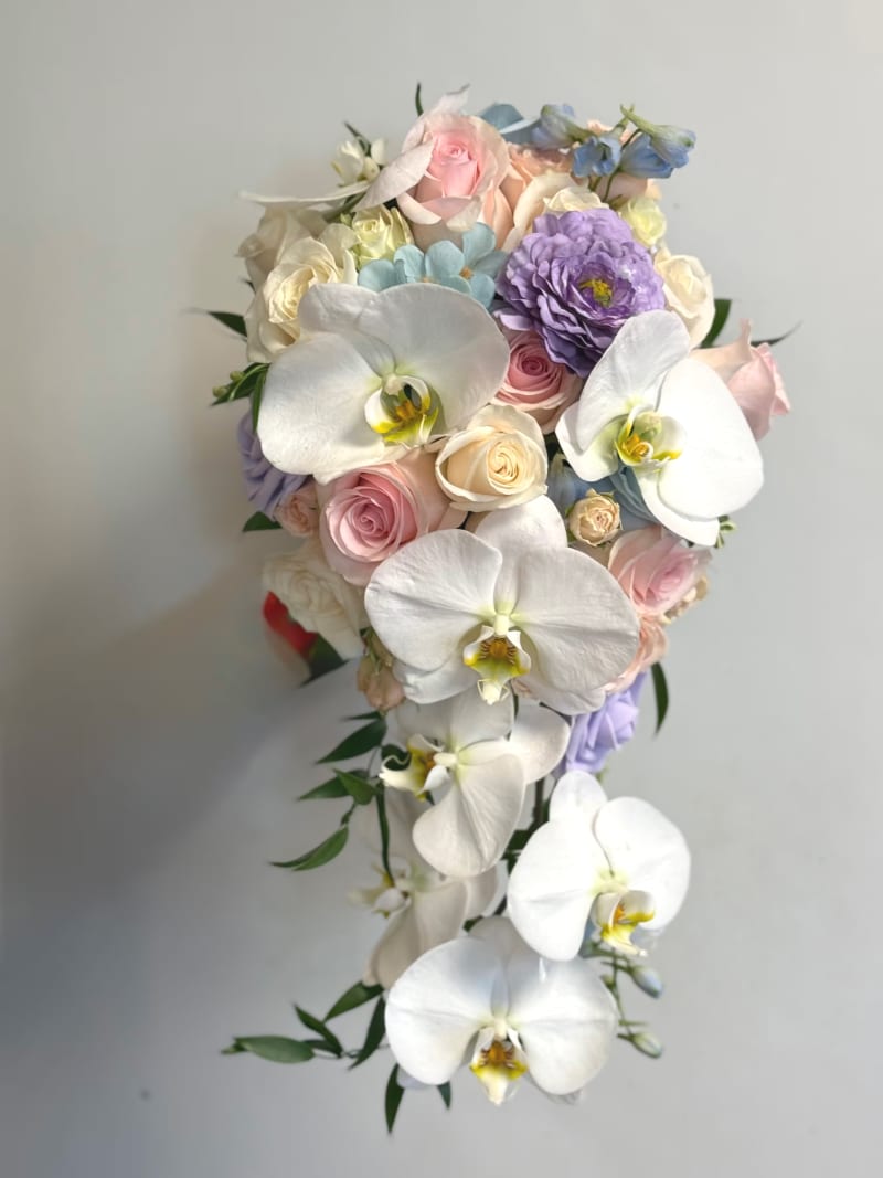 Pastel cascade bridal bouquet by Liz’s Flowers with white orchids, blush roses, lavender flowers, and soft blue accents.