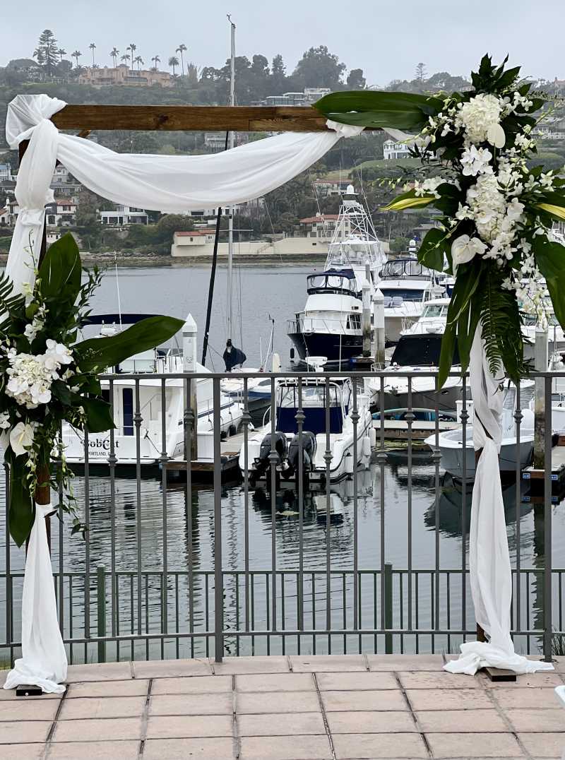 Wooden ceremony arch decorated with white hydrangeas, orchids, anthuriums, tropical greenery, and flowing white fabric overlooking the marina at Kona Kai in San Diego.
