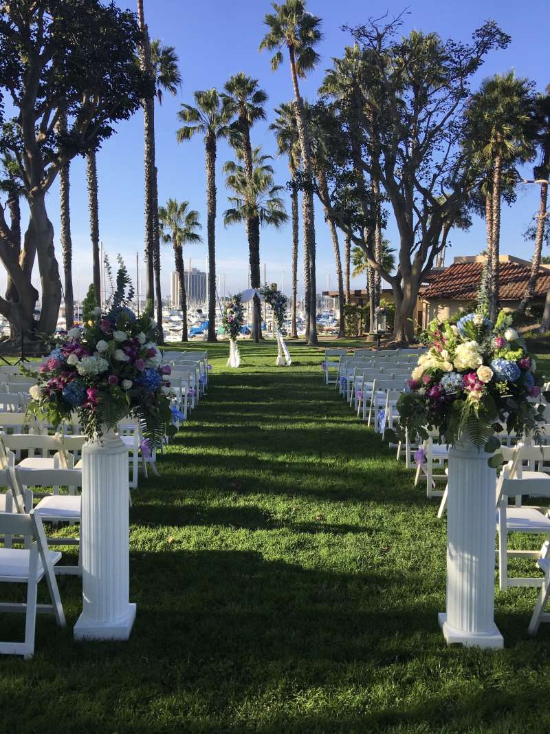 Garden wedding ceremony on the North Lawn at Marina Village featuring rows of white chairs, colorful floral arrangements on pedestals, and a floral arch framed by palm trees and marina views.