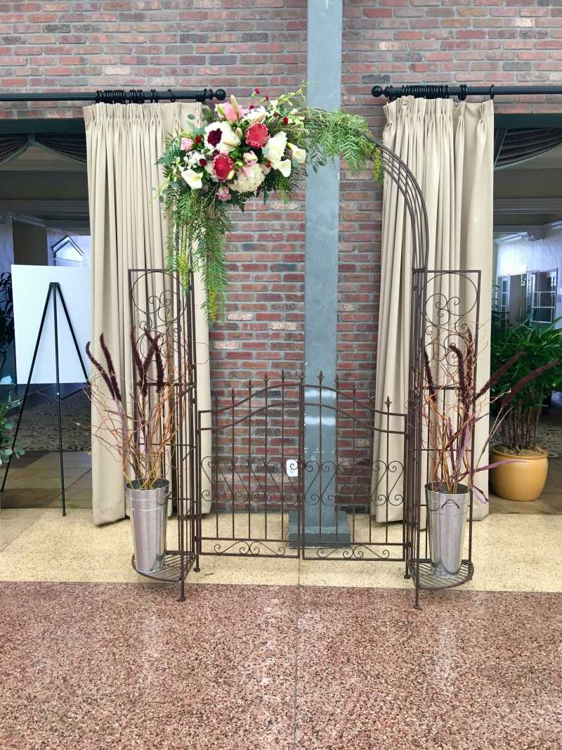 Vintage-style metal garden gate arch decorated with pink, burgundy, and white flowers, cascading greenery, and tall branch arrangements for a wedding backdrop or entrance display.