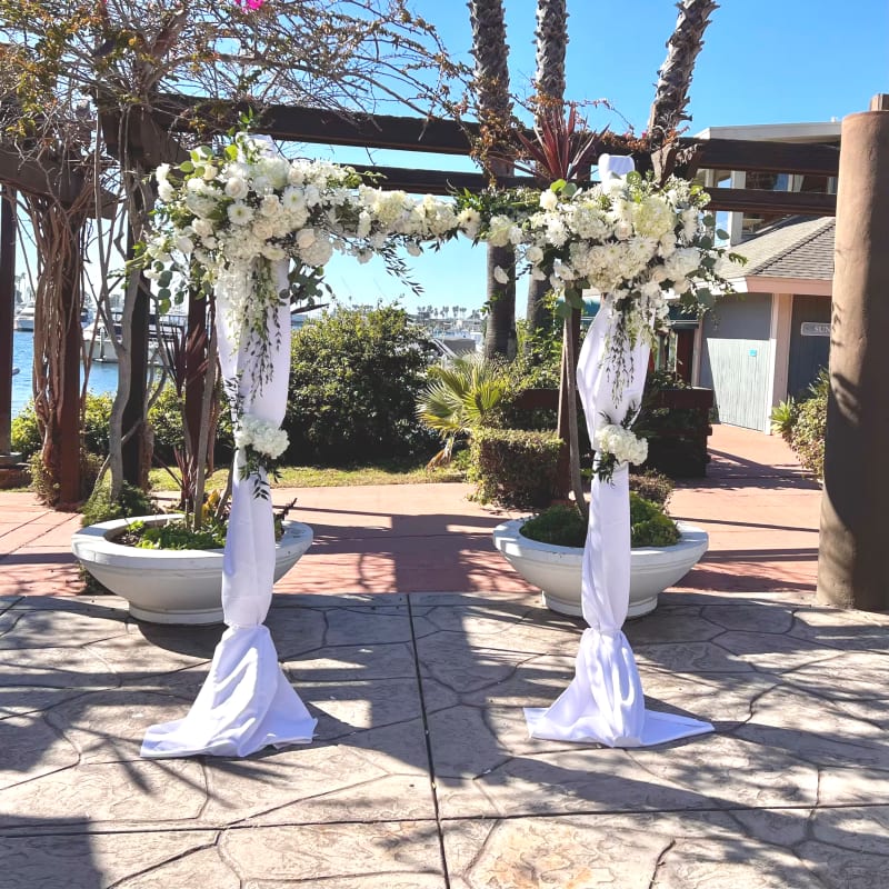 White wedding ceremony arch with lush white flowers and draped fabric at a waterfront outdoor in Marina Village