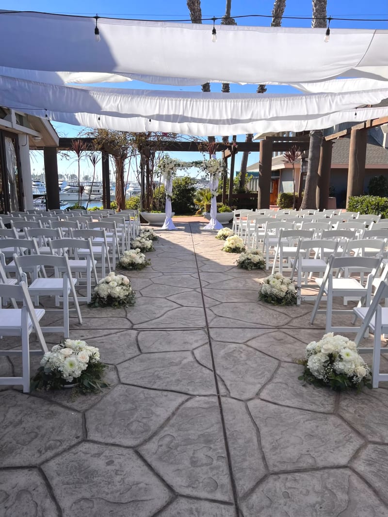 Outdoor wedding ceremony setup with white chairs, a floral arch, and repurposed white centerpieces arranged along the aisle at Marina Village Conference Center