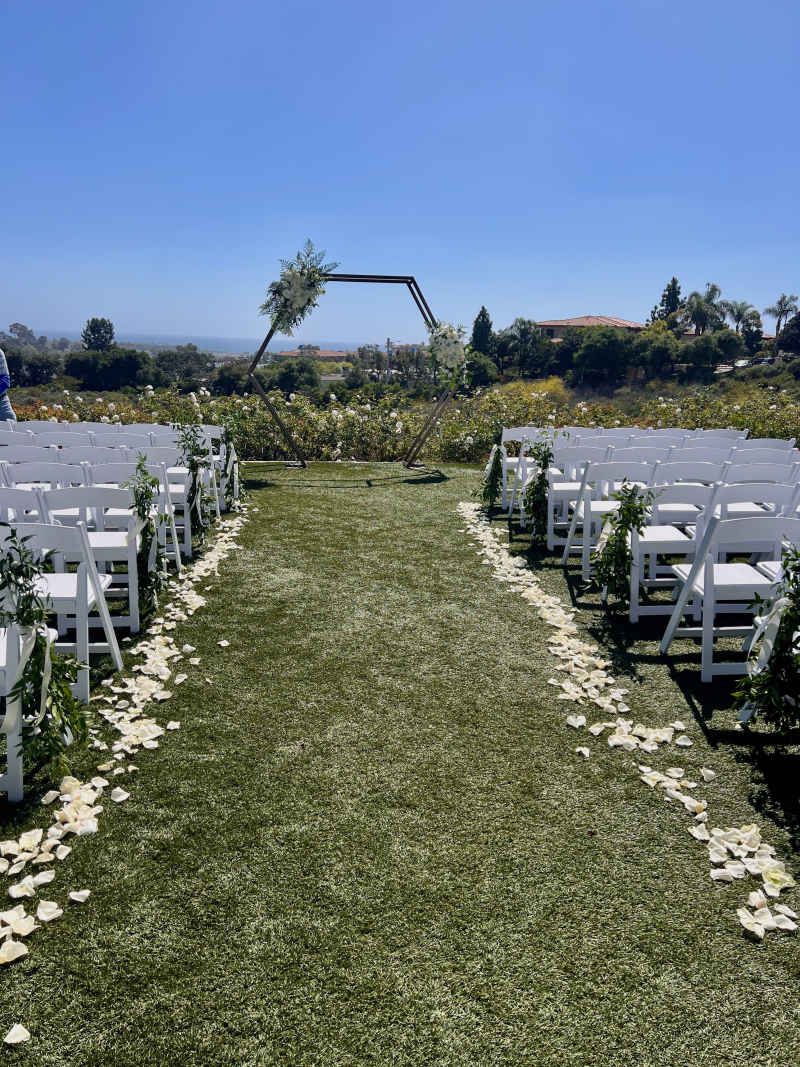 Wedding ceremony setup at The Crossings at Carlsbad featuring rows of white chairs, greenery-tied aisle chairs, white rose petals, and a geometric hexagon arch with white floral accents.