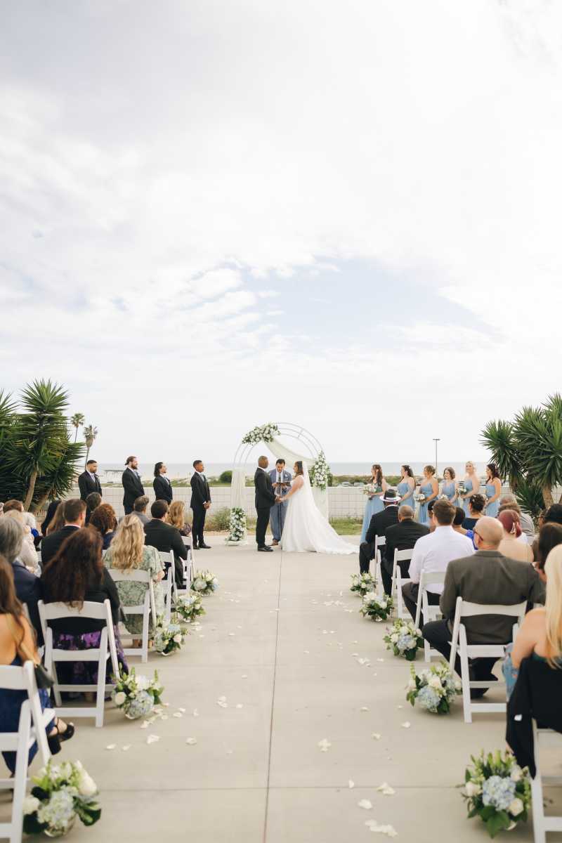Outdoor wedding ceremony featuring white chairs, scattered rose petals, and repurposed reception centerpieces arranged along the aisle leading to a white floral arch.