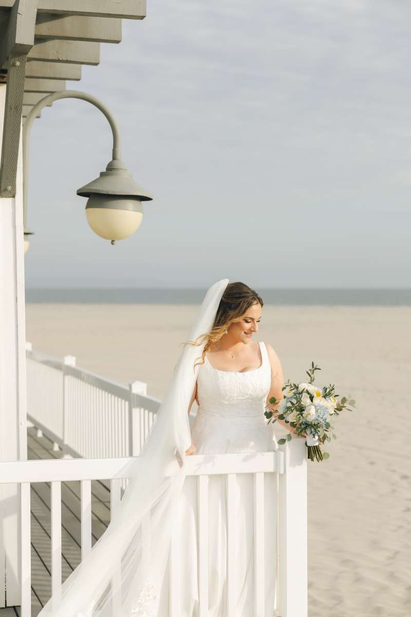 Bride wearing a white wedding dress and veil, holding a bouquet of blue hydrangeas, white roses, and greenery while standing on a beachside boardwalk.