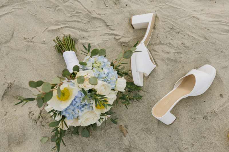 Bridal bouquet with blue hydrangeas, white roses, white blooms, and greenery styled on the sand beside white bridal shoes for a beach wedding.