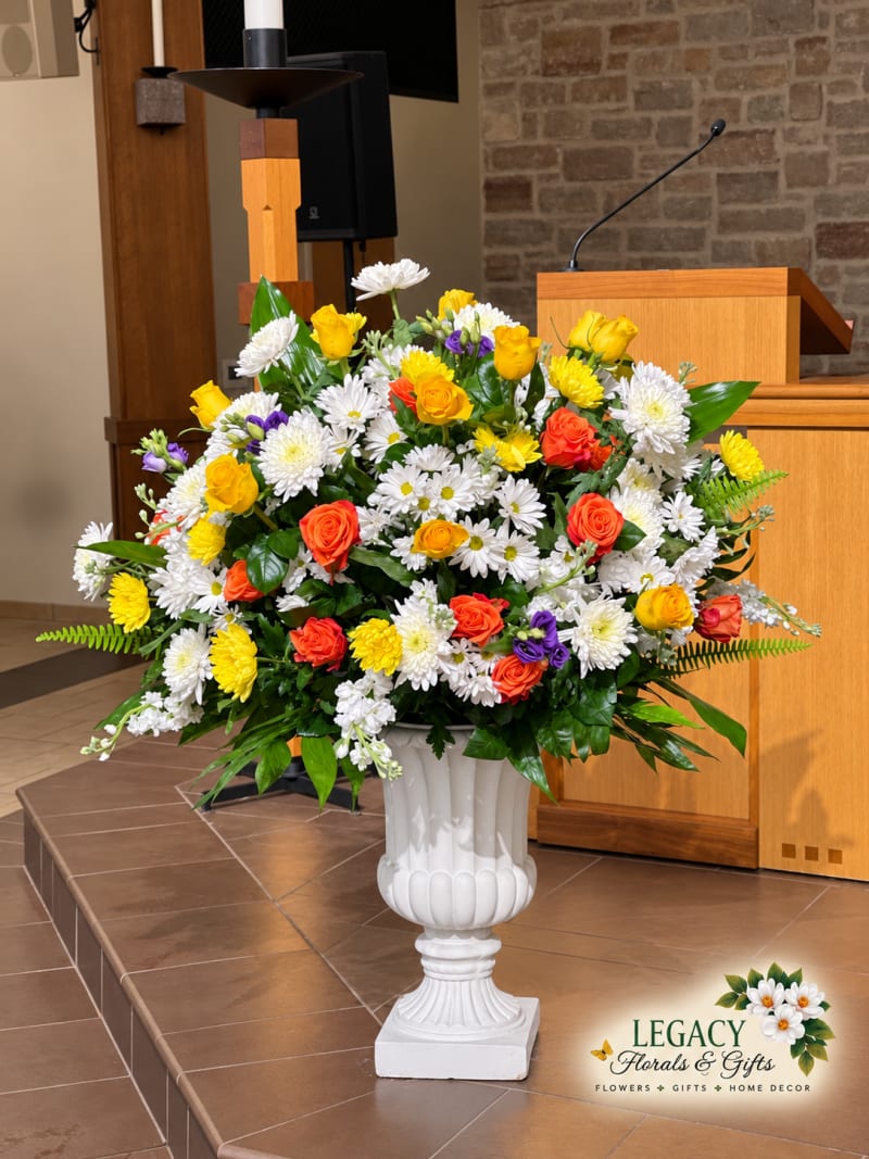 Yellow and orange roses with white flowers, purple accents, and greenery in a white pedestal urn beside a wooden church pulpit.