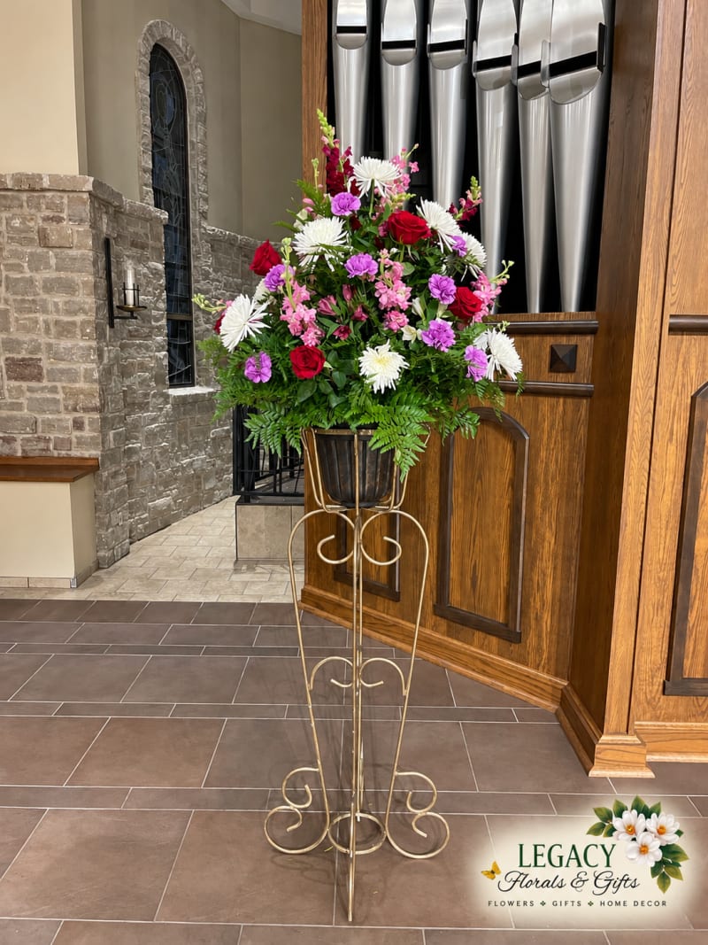 Pink, purple, and white floral arrangement on a tall gold scrollwork stand beside wooden organ pipes and a stone church wall.