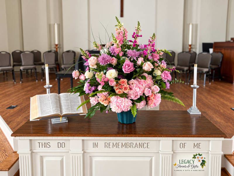 Pink and lavender floral arrangement in a teal container on a white communion table beside an open Bible and tall candlesticks.
