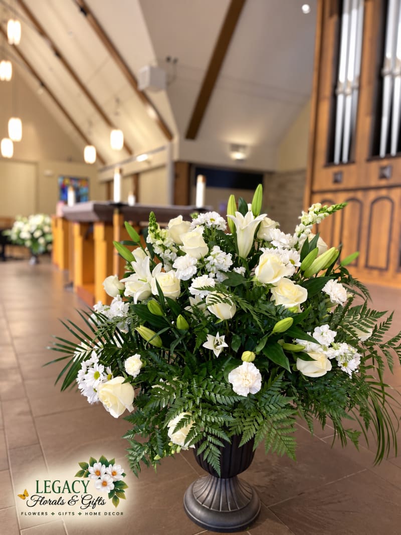 White roses and lilies with lush greenery in a dark pedestal urn inside a church, with organ pipes in the background.
