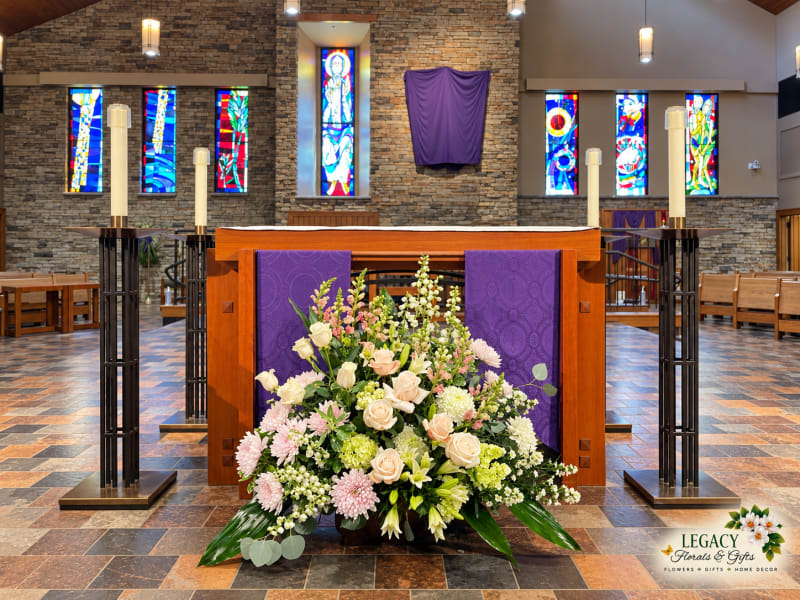 Low arrangement of cream roses, pale pink flowers, and greenery before a wooden church altar with purple panels and stained-glass windows.