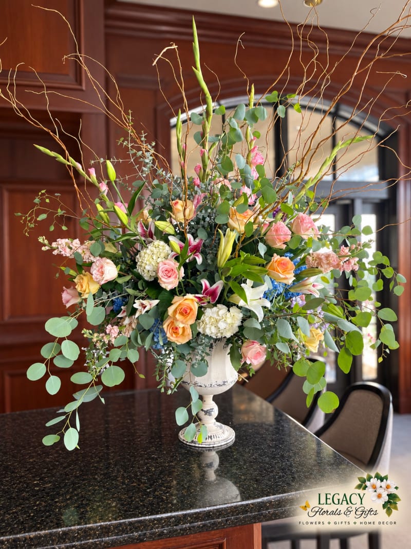 Pastel roses, lilies, and hydrangeas with cascading eucalyptus and tall curly branches in a distressed white pedestal urn on a dark granite counter.