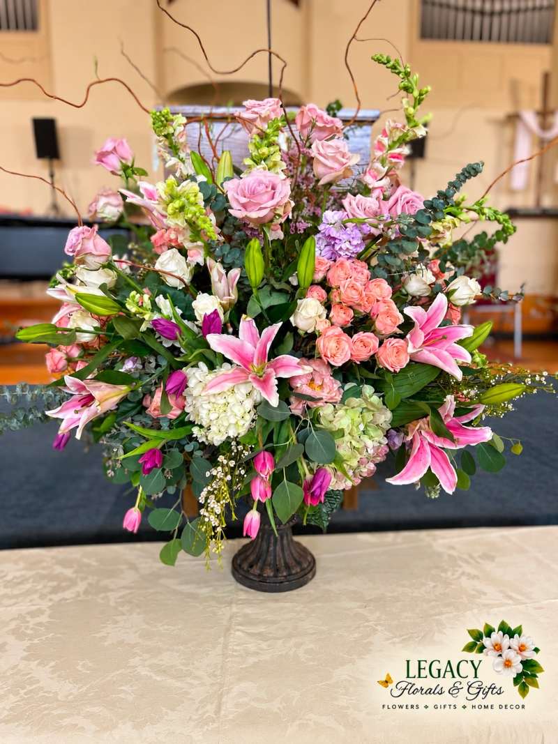 Grand Pink lilies and roses with white hydrangeas, greenery, and tall branches in a dark pedestal urn.