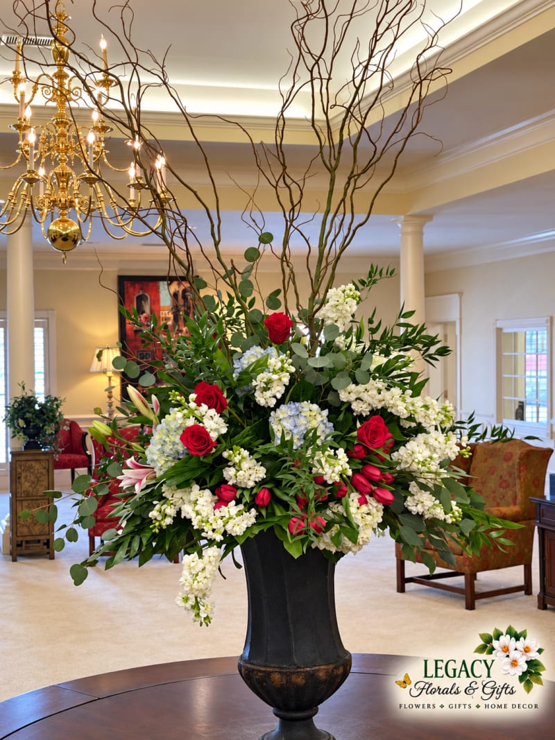 Red and white floral arrangement with cascading greenery and in a dark urn on a round wooden reception table.
