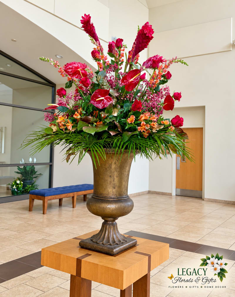 Tall red and orange floral arrangement with lush greenery in a bronze pedestal urn on a wooden stand in a church lobby.